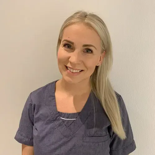 Kvinne med blondt hår iført en blå skrubbtopp, smilende. Hvit vegg i bakgrunnen.