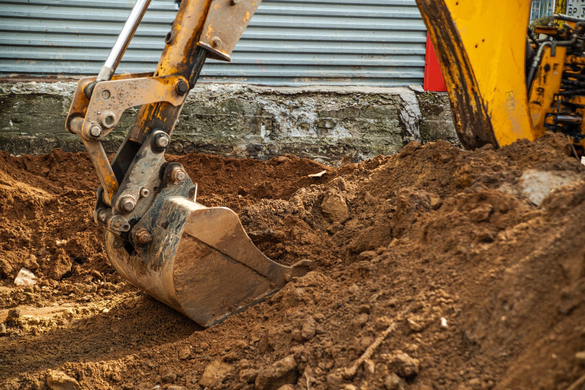 Excavator bucket digging dirt at a construction site beside a corrugated metal wall