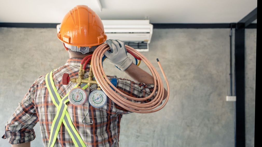 Worker in orange hard hat carrying coiled cables and standing by a wall-mounted unit in a room