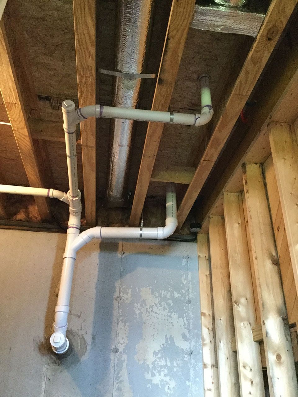 Exposed white plumbing pipes beneath wooden ceiling joists in an unfinished basement ceiling