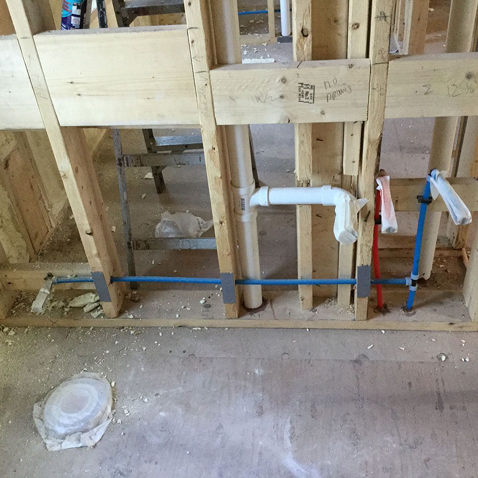 Exposed bathroom plumbing in framed wall with blue PEX pipes and PVC drain lines under construction