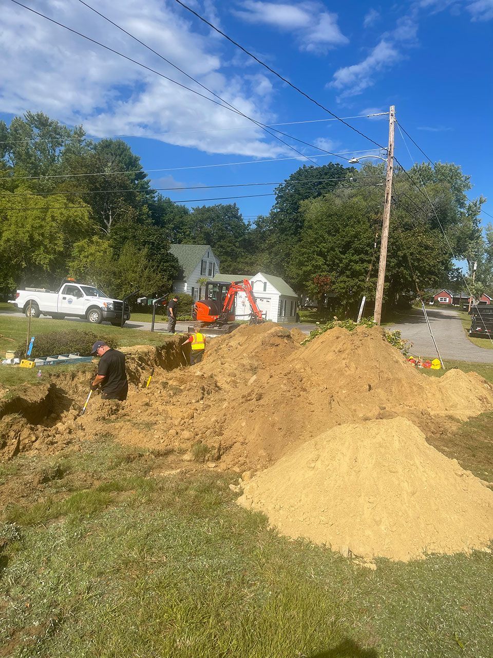 Construction site with dirt piles, utility pole, white truck, and workers near a house under a blue sky