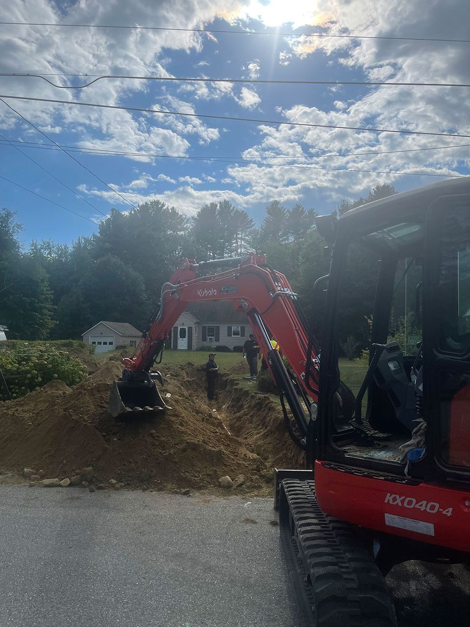 Red excavator digging a dirt mound beside a road under a cloudy blue sky
