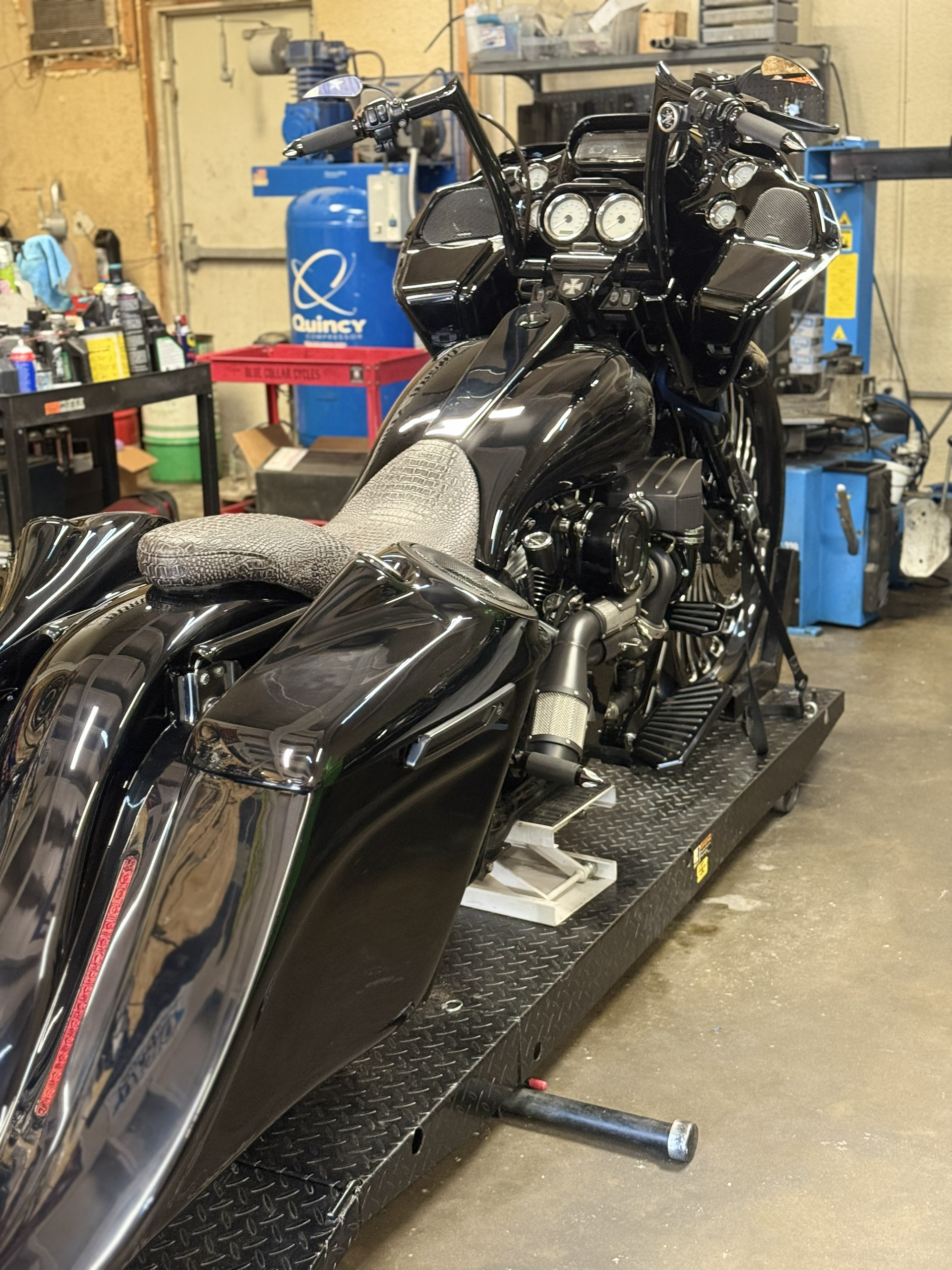 Black custom motorcycle in a workshop, seen from the rear with chrome details and tool benches in the background