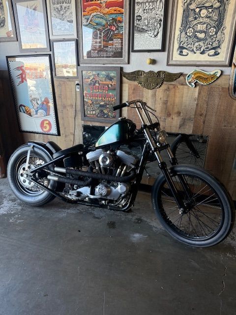 A custom motorcycle with a dark green tank parked in a room with vintage posters on a wood-paneled wall.