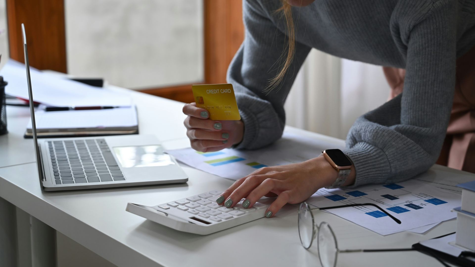 Person uses a calculator while holding a gold credit card near a laptop and financial documents on a desk.