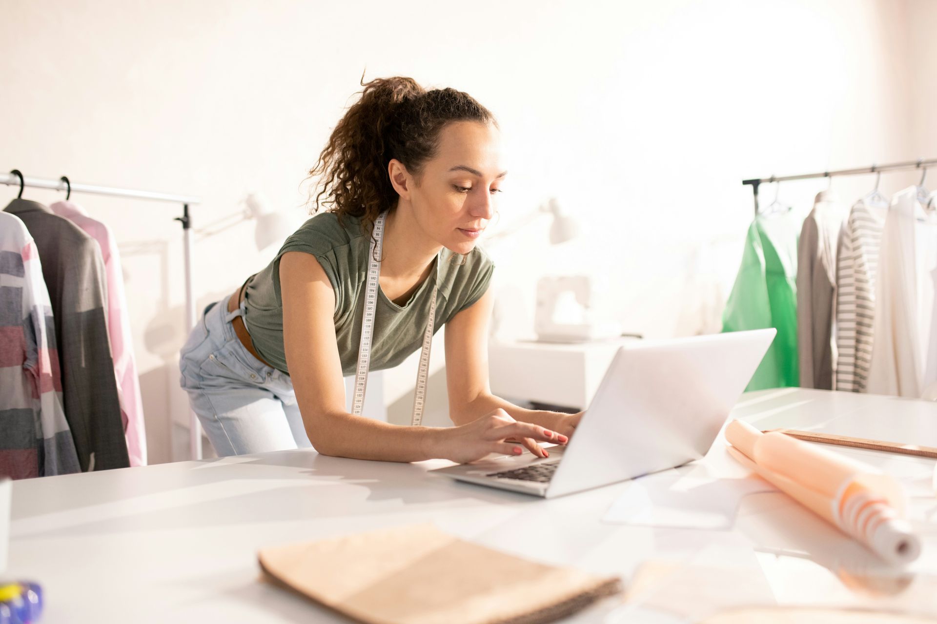 Woman leans over a laptop at a white table in a workshop, working with fabric and sewing supplies.