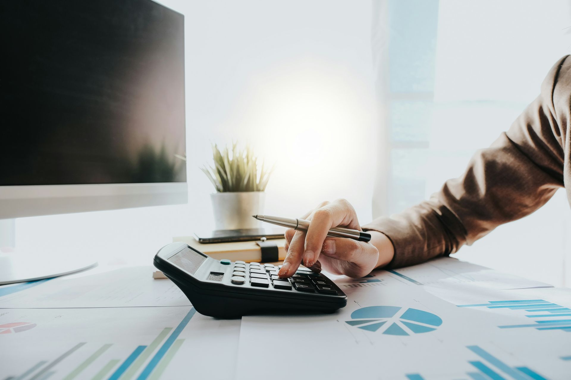 Person using a calculator with a pen, examining financial documents on a desk, next to a computer.