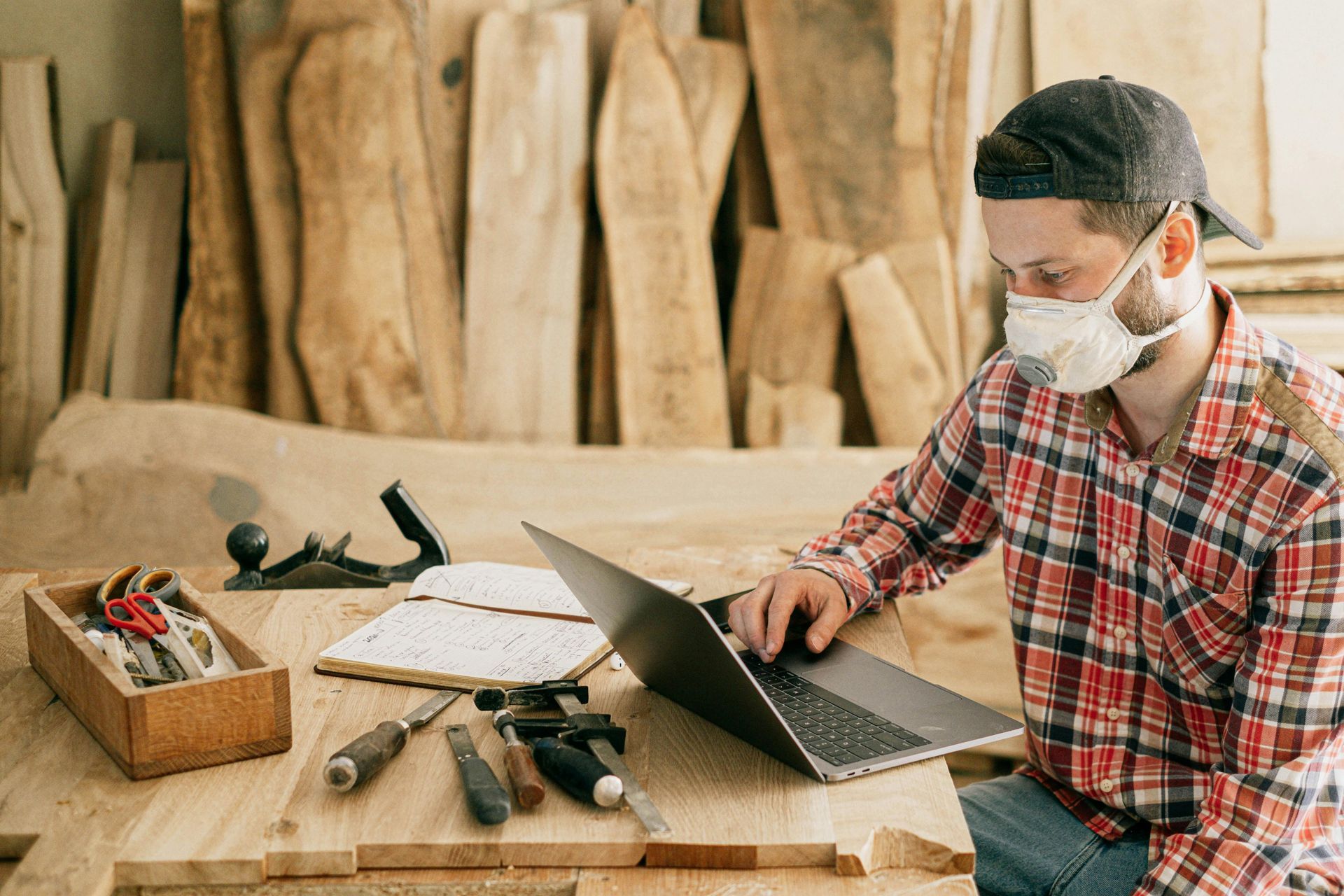 A person in a workshop wears a mask and cap, working on a laptop with tools nearby.