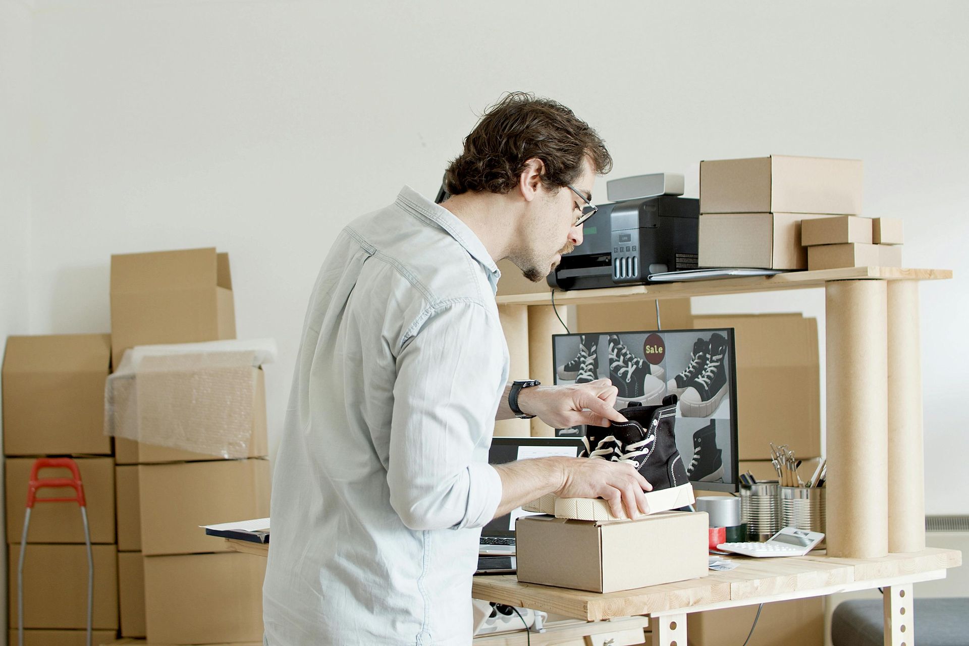 Man packing a box at a desk with computer and other boxes, light-colored room.