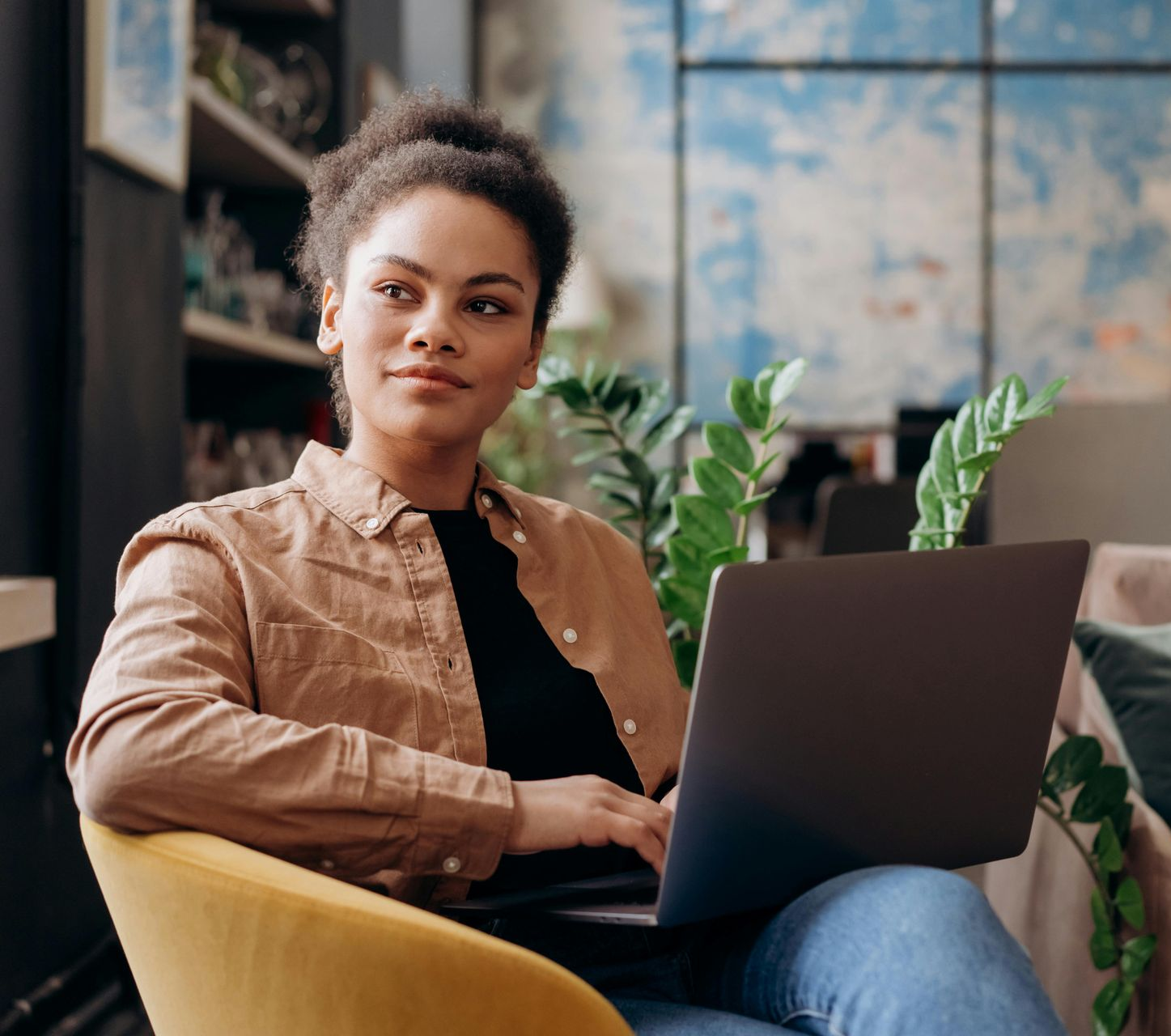 Woman in a brown shirt and jeans sits in a yellow chair with a laptop, looking thoughtful.
