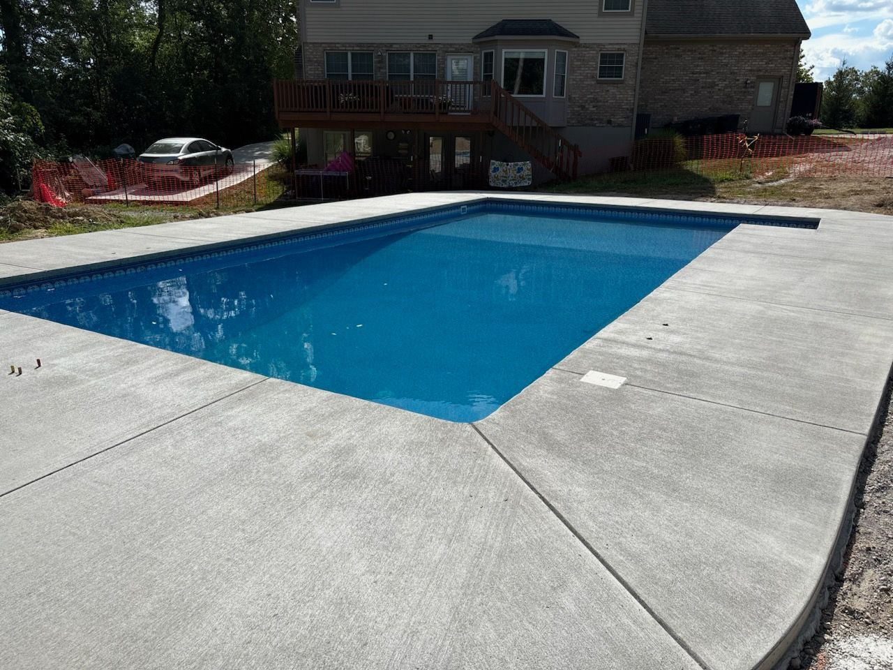 A bright blue rectangular swimming pool surrounded by a light grey concrete patio in a grassy backyard near a house.