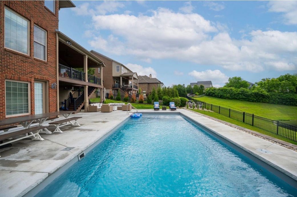 A long rectangular swimming pool with blue water sits on a concrete patio next to a multi-story brick house.