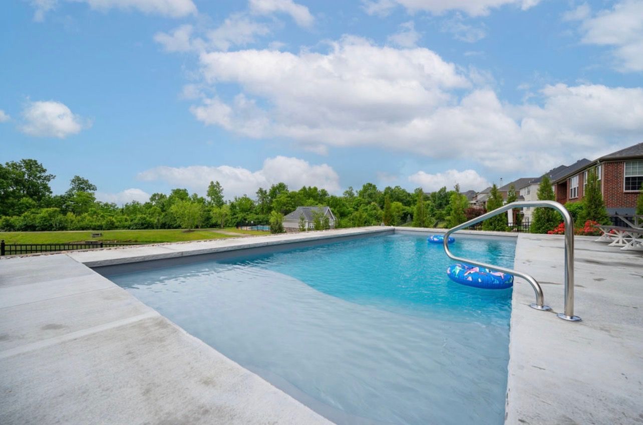 A blue rectangular swimming pool with a shallow tanning ledge, featuring a metal handrail and blue floats under a blue sky.