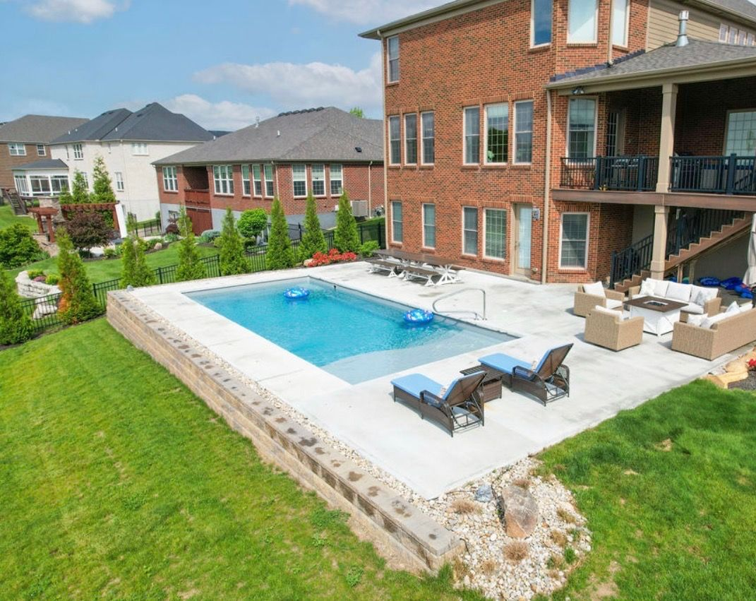 An elevated backyard swimming pool with a concrete patio, lounge chairs, and outdoor seating next to a brick house.