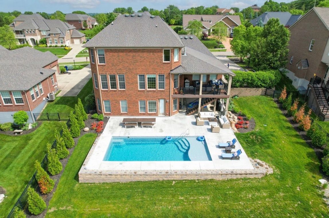 Aerial view of a brick house with a backyard pool, patio furniture, and landscaped lawn in a suburban neighborhood.