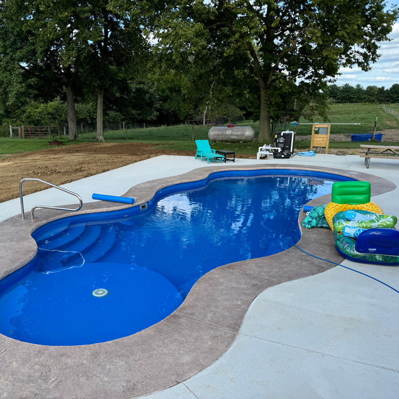 A blue, free-form swimming pool with steps, surrounded by a light-colored concrete deck in a backyard with trees.