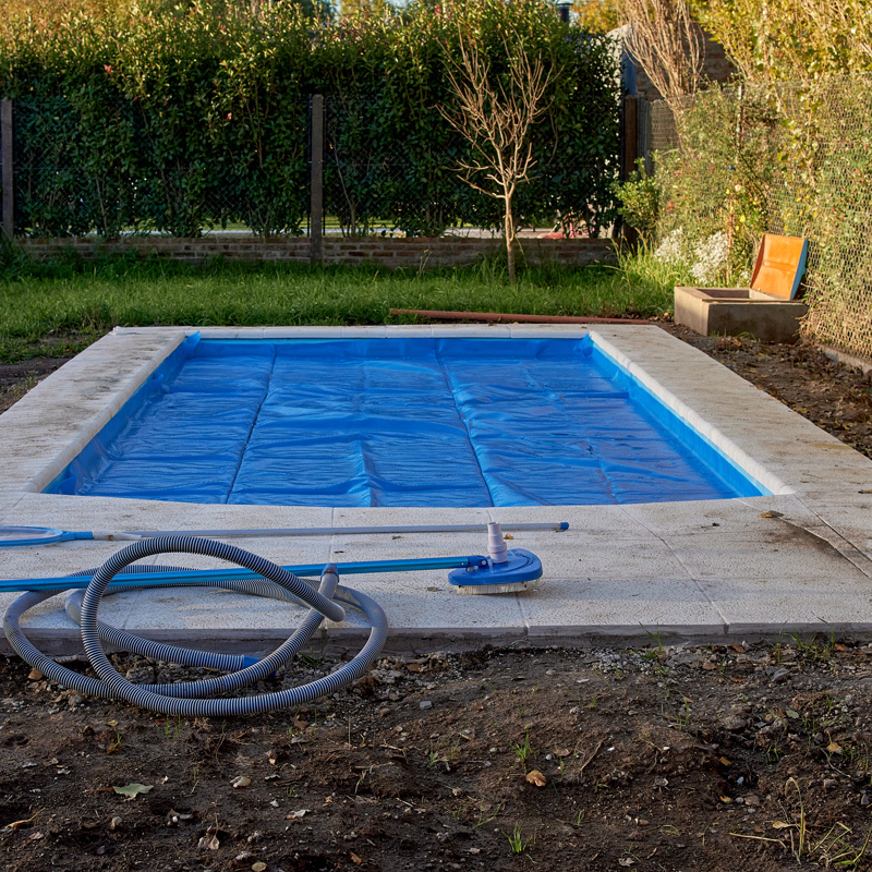 A blue pool cover on a backyard rectangular pool, with cleaning equipment resting on the light stone decking.
