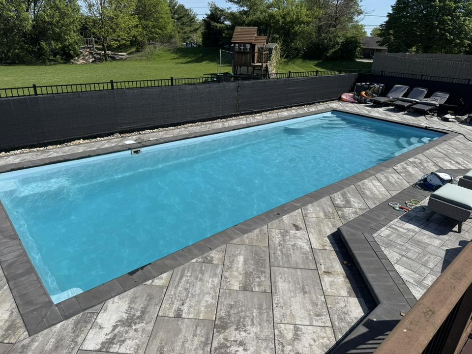 A rectangular, light blue swimming pool surrounded by gray stone patio tiles and a black privacy fence in a backyard.