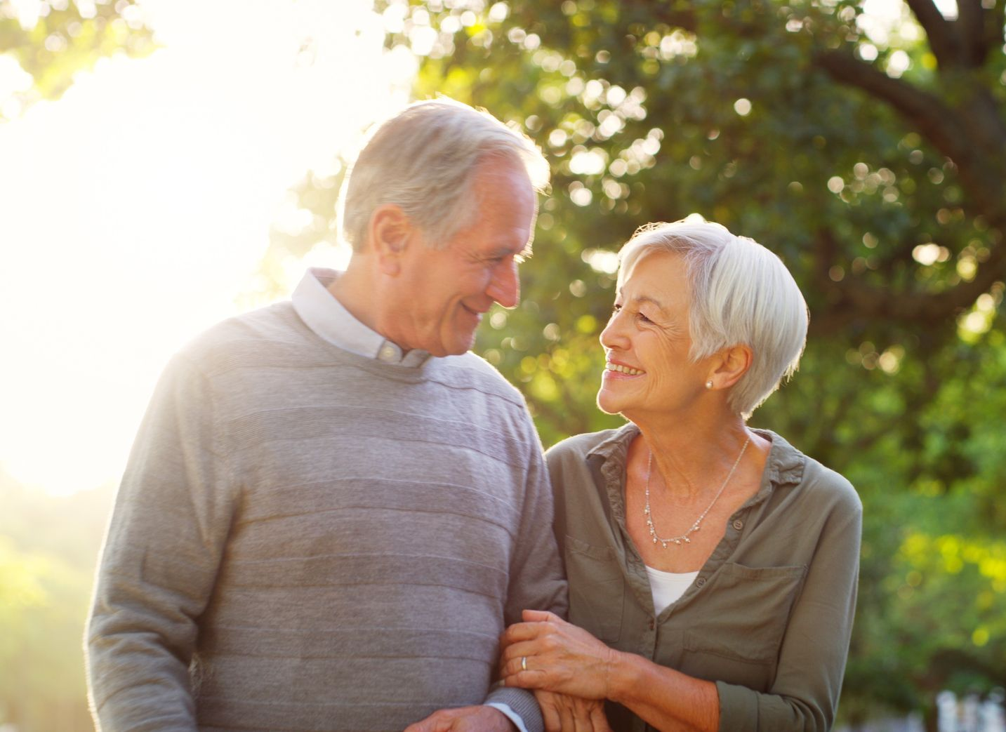 Two smiling seniors standing together outdoors in warm sunlight, holding hands and looking at each other.