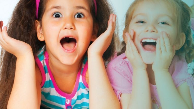 Two girls in colorful shirts looking at the camera with a surprised look on their faces. Their hands are by their chins.