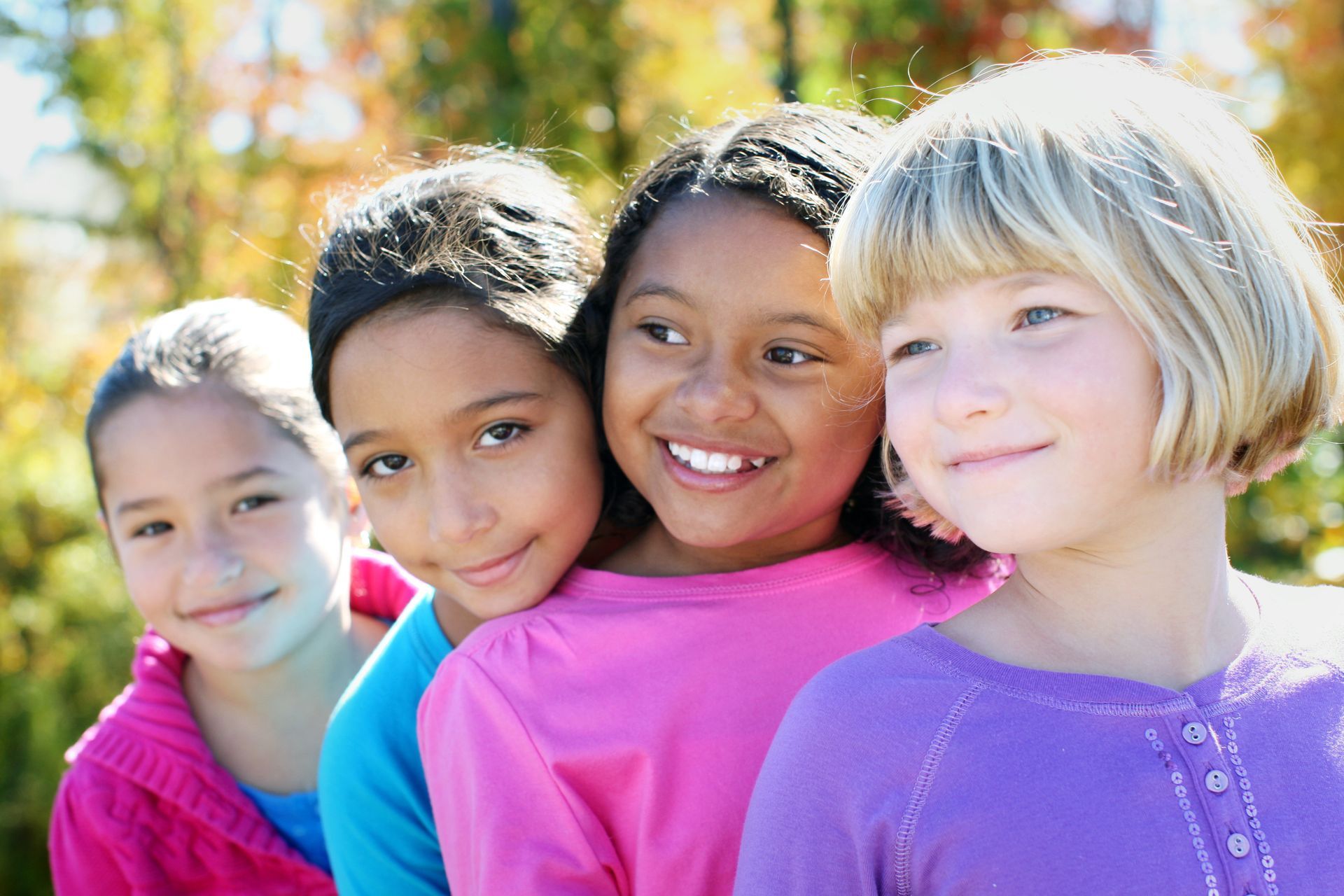 Young girls standing in a line in a park. They are all smiling and wearing bright tops.