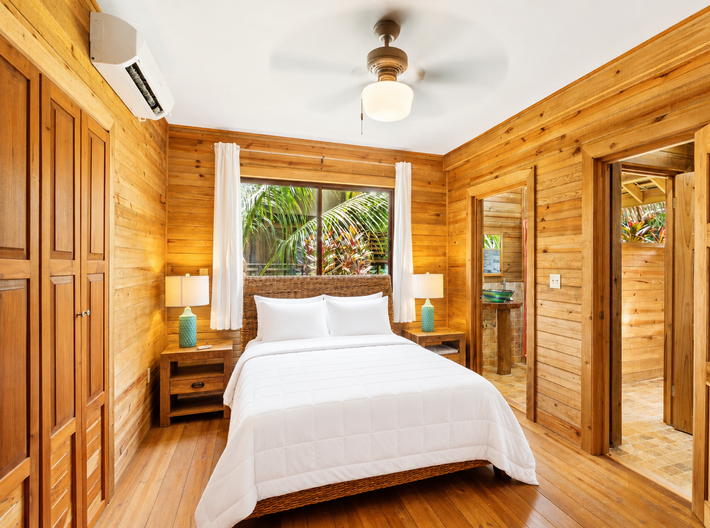 Wood-paneled bedroom with a white bed, ceiling fan, and large windows overlooking tropical greenery