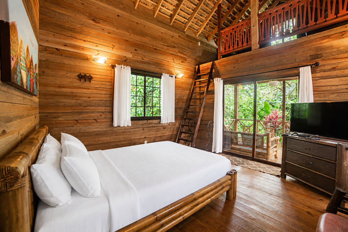 Rustic wooden bedroom with a large bed, window, TV, and balcony doors overlooking greenery