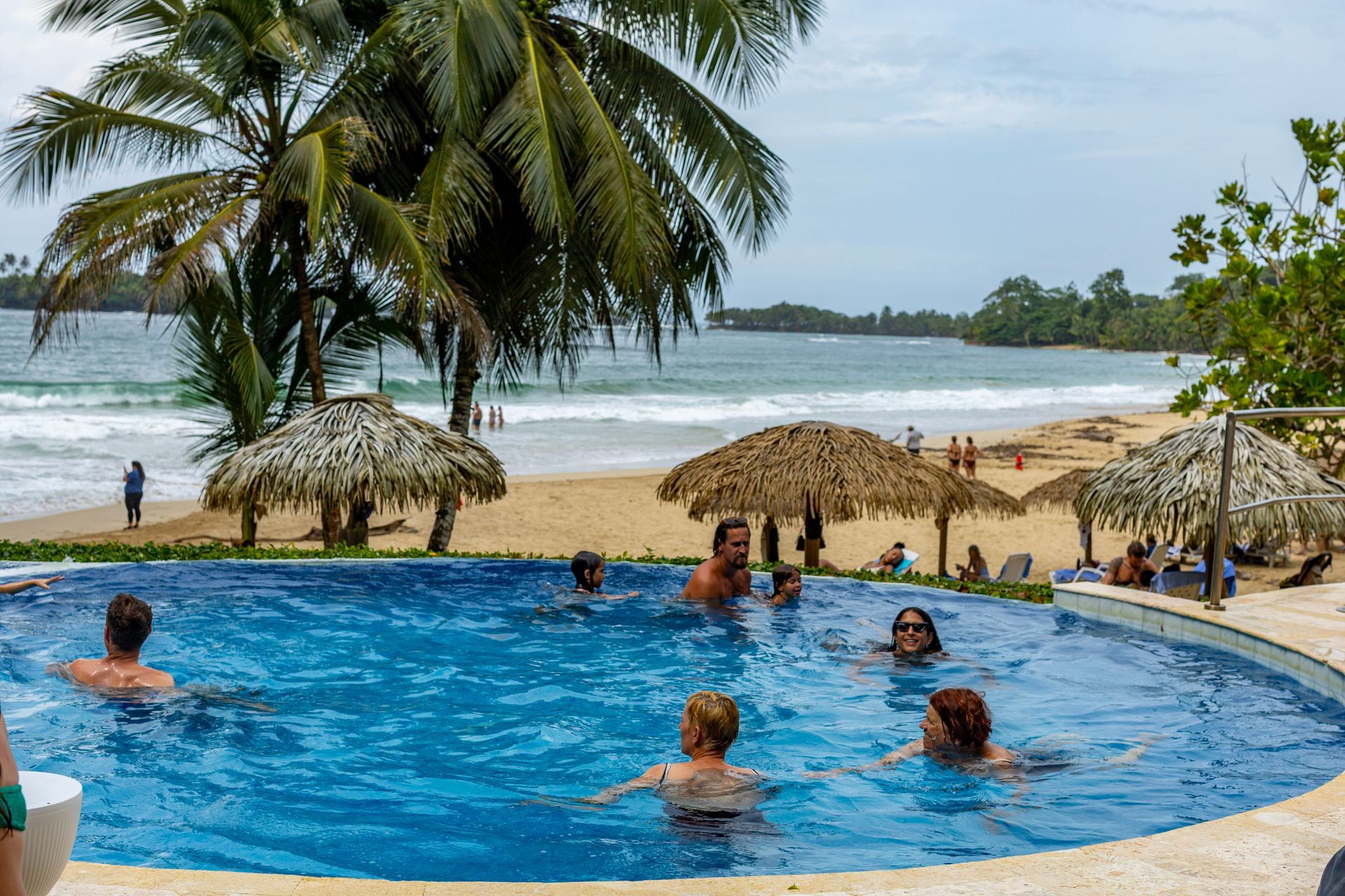 People swimming in a blue pool beside a sandy beach with palm trees and straw umbrellas