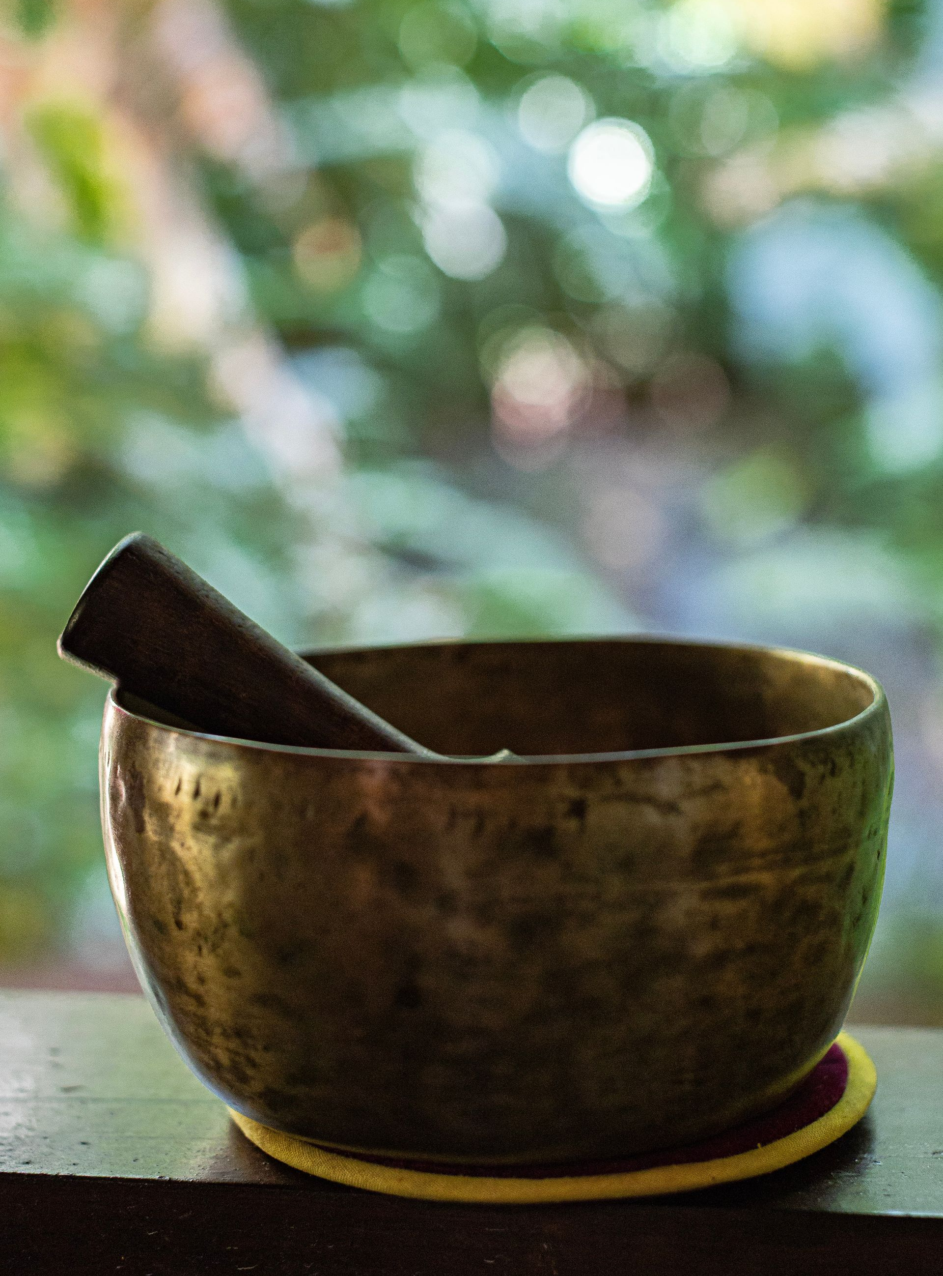 Bronze singing bowl with wooden striker on a table, against a blurred green background