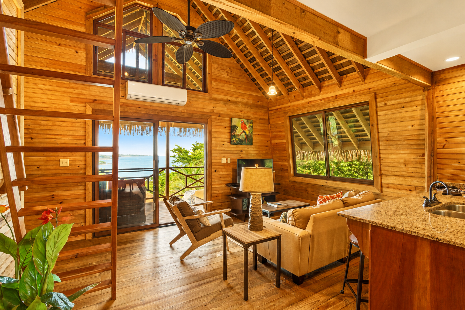 Wooden cabin interior with vaulted ceiling, lounge chairs, kitchen counter, and ocean view through glass doors