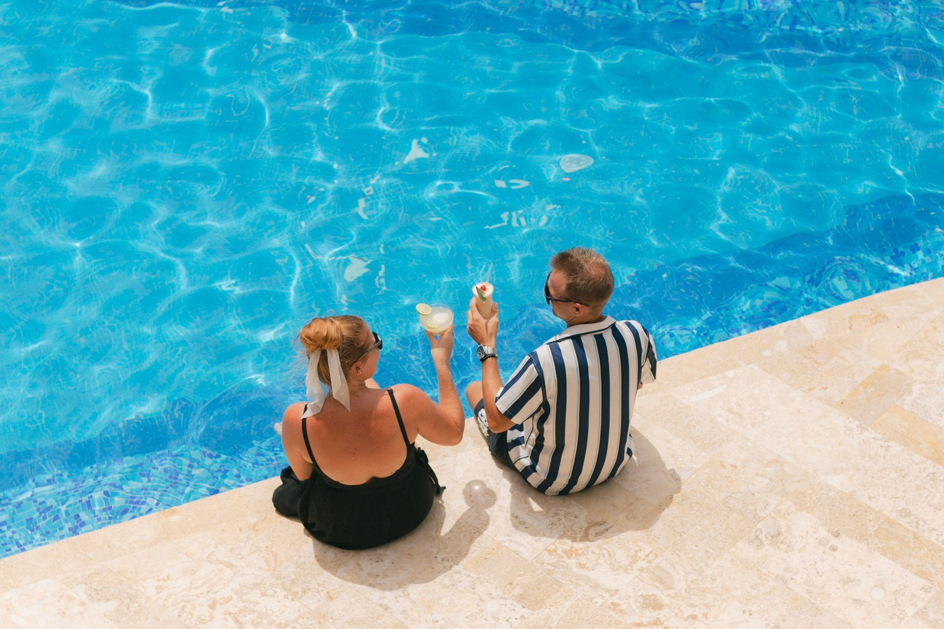 Two people sit by a bright blue pool, dipping their feet in the water.
