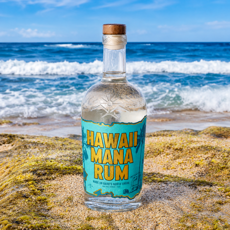 Hawaii Mana Rum bottle on a coral rock at the shoreline on Oahu's North Shore, turquoise label facing the surf, handcrafted by Haleiwa Distilling Company.