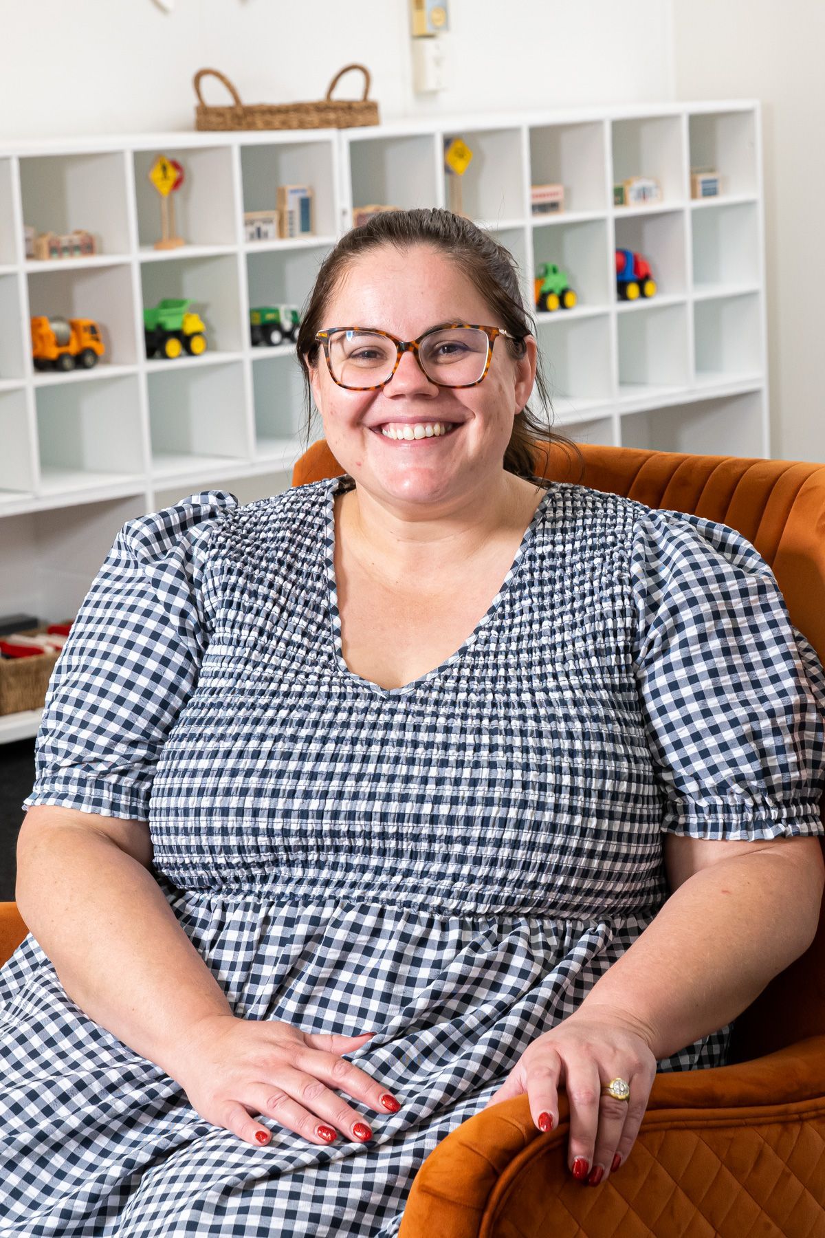Director if Childcare wearing glasses and a plaid dress sits in an orange chair