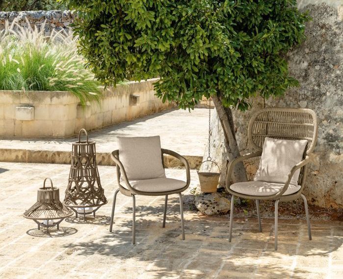 Two outdoor chairs with cushions near a tree, stone wall in background, lanterns on the ground.