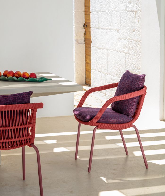 Two outdoor dining chairs, red woven backs and seats, purple cushions, by a table.