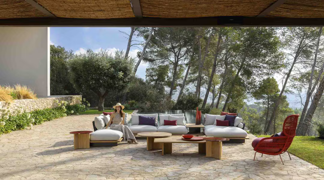 Outdoor seating area with woman sitting, white couches, red chair, trees, and stone patio.