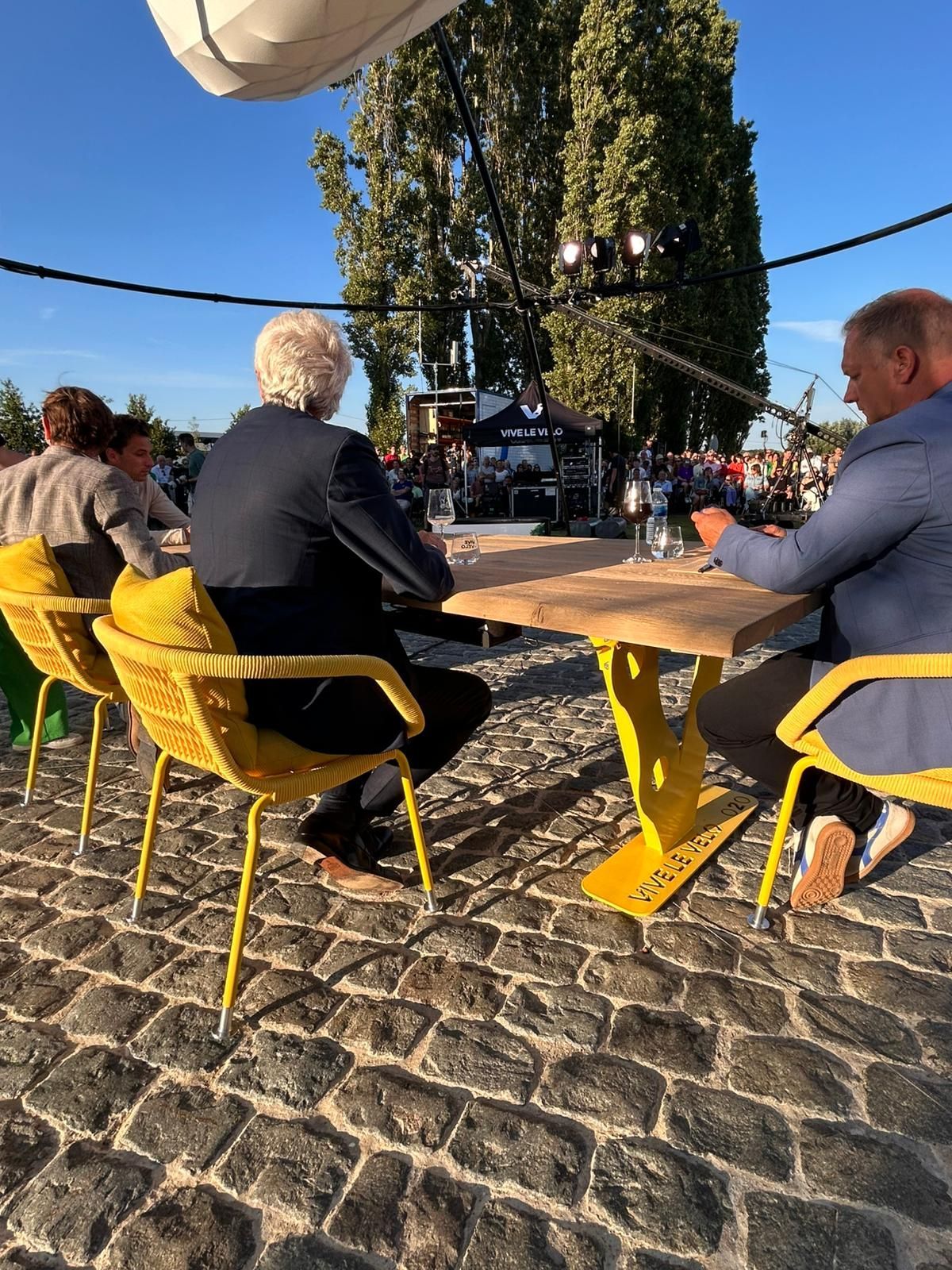 People seated at a yellow table, outdoor setting, cobblestone ground.