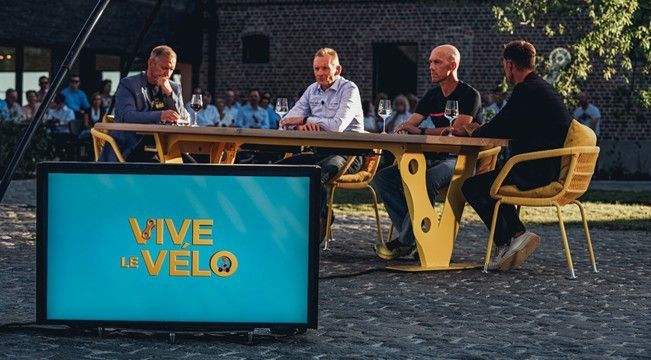 Four men at a yellow table, a show taping outdoors. A screen displays the show's logo. Guests in audience.