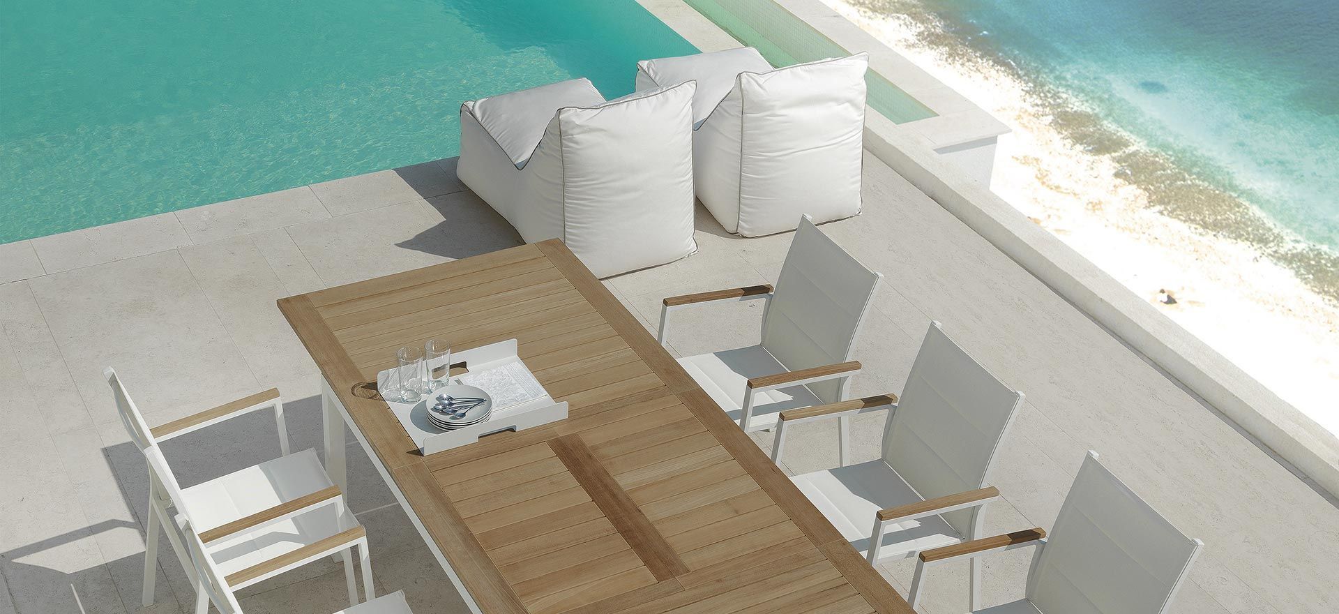 Outdoor dining set near a pool and beach. White chairs, wooden table, and white bean bag chairs are visible.