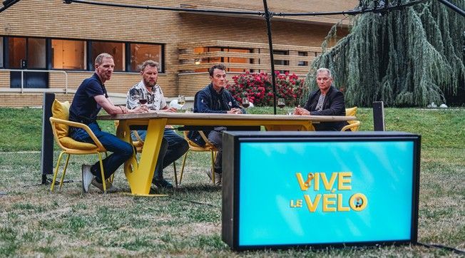 Four men sit at a yellow table outside. A lighted sign in front reads 