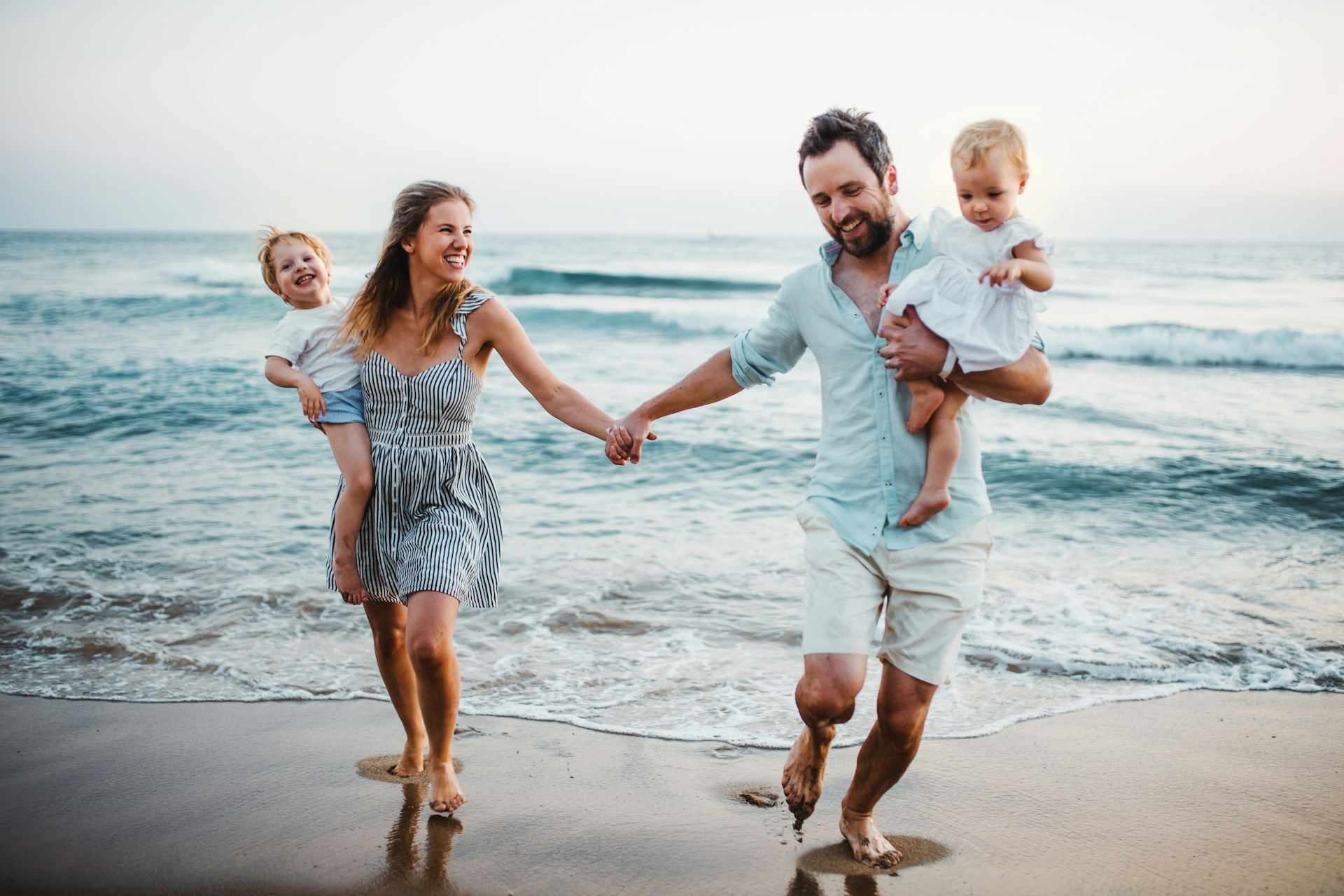 A family of four walking and smiling on a beach. Parents holding young children as they walk along the shore.
