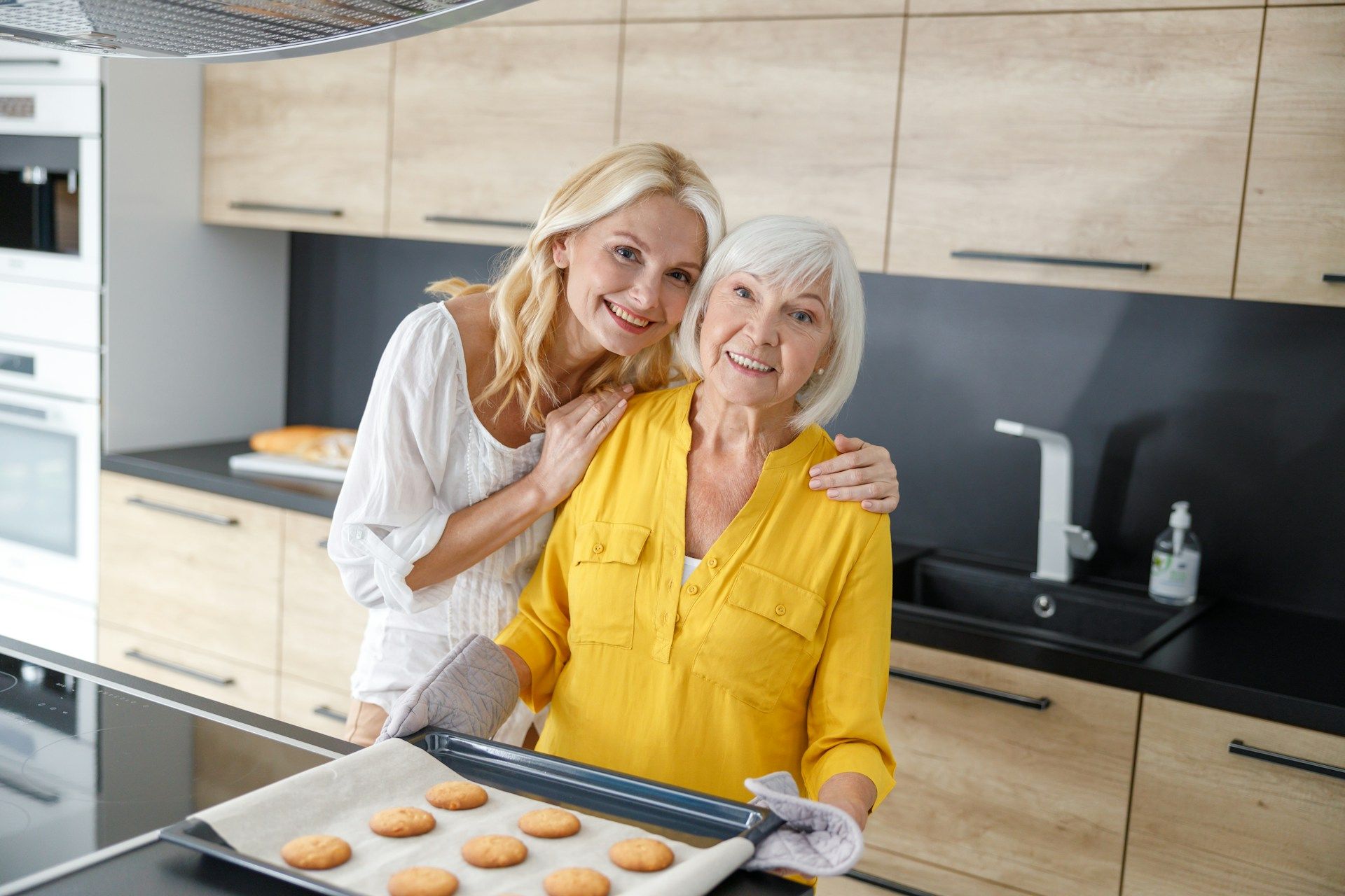 Woman hugs older woman holding a tray of cookies in a kitchen. Both smile at the camera.
