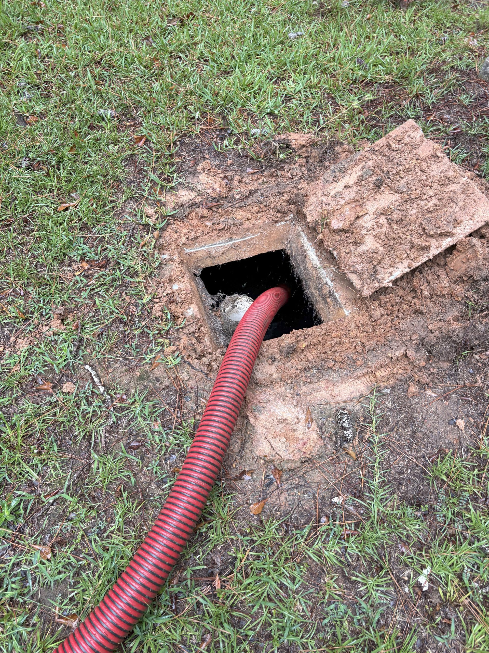 Red corrugated hose entering a small square hole in the ground beside a grassy patch and concrete cover