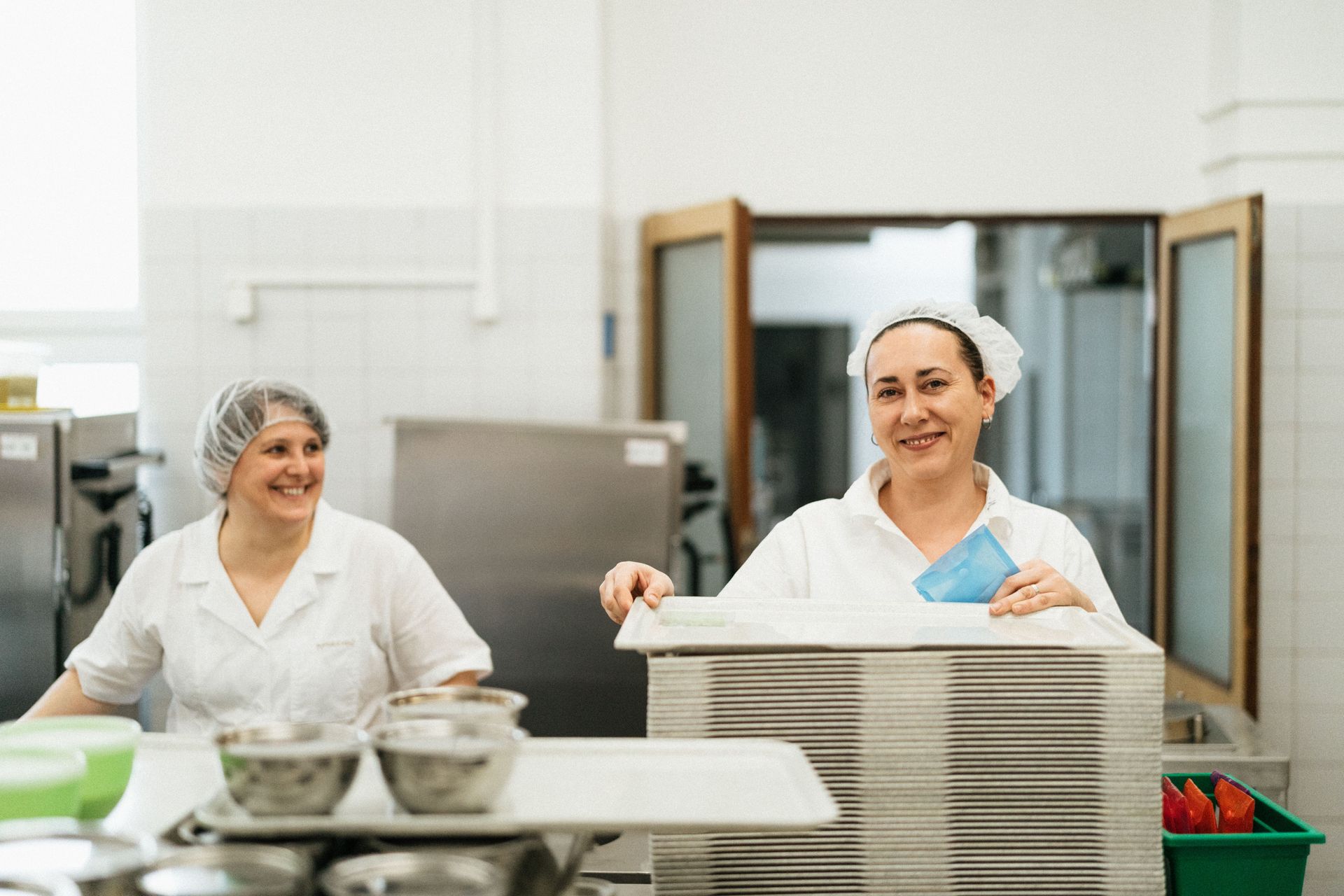 Two workers in a bright kitchen stack trays beside stainless steel tables and bowls