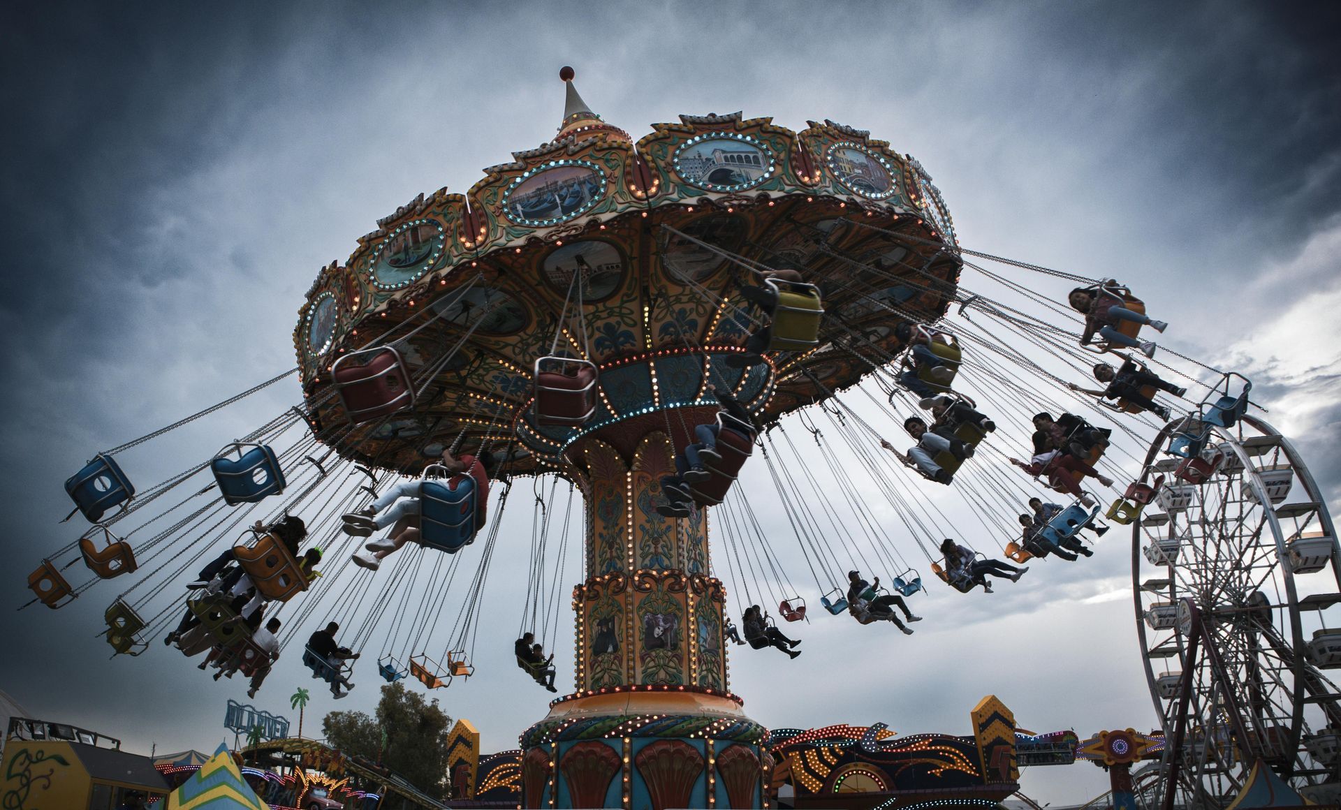 Carousel swing ride at a fair, with people in the seats. A Ferris wheel is in the background under a cloudy sky.