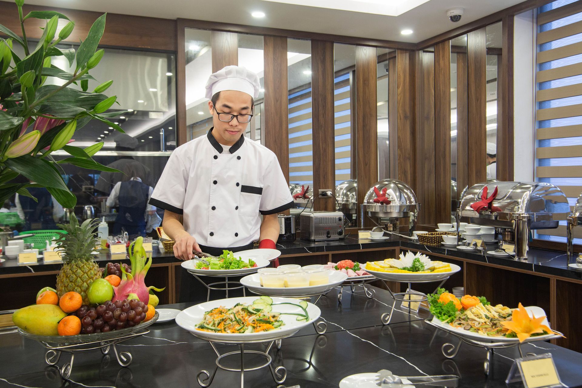 Chef preparing food at a buffet counter with platters of colorful dishes and fruit in a bright dining room