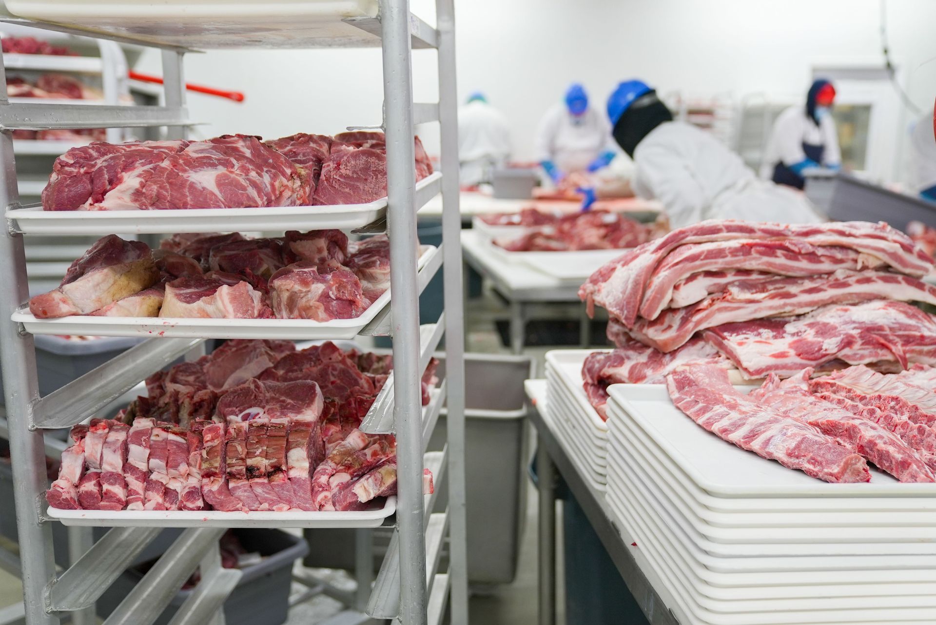 Meat trays on racks in a food processing room, with workers sorting meat in the background