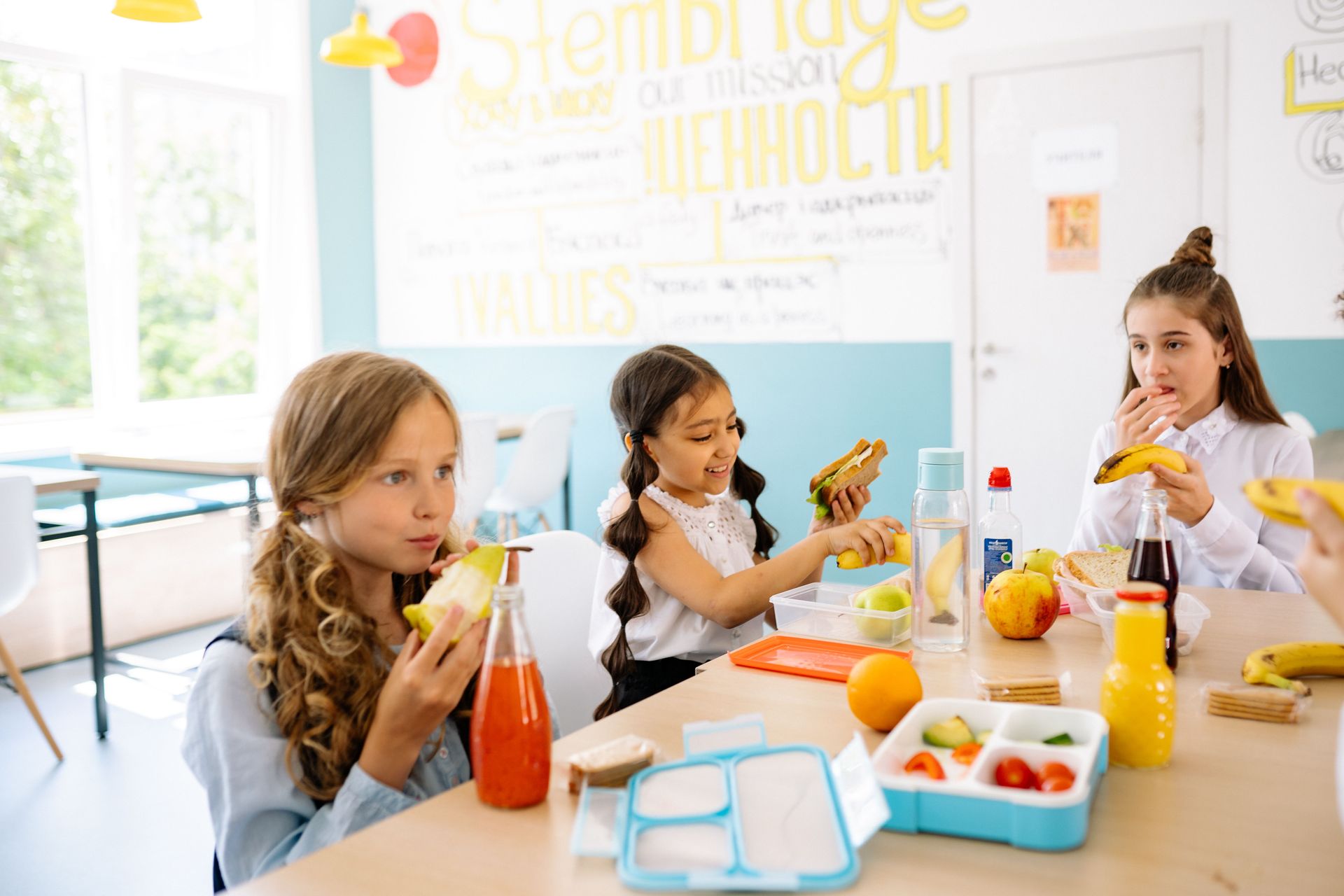 Three children sitting at a table, eating snacks and drinking juice in a bright classroom