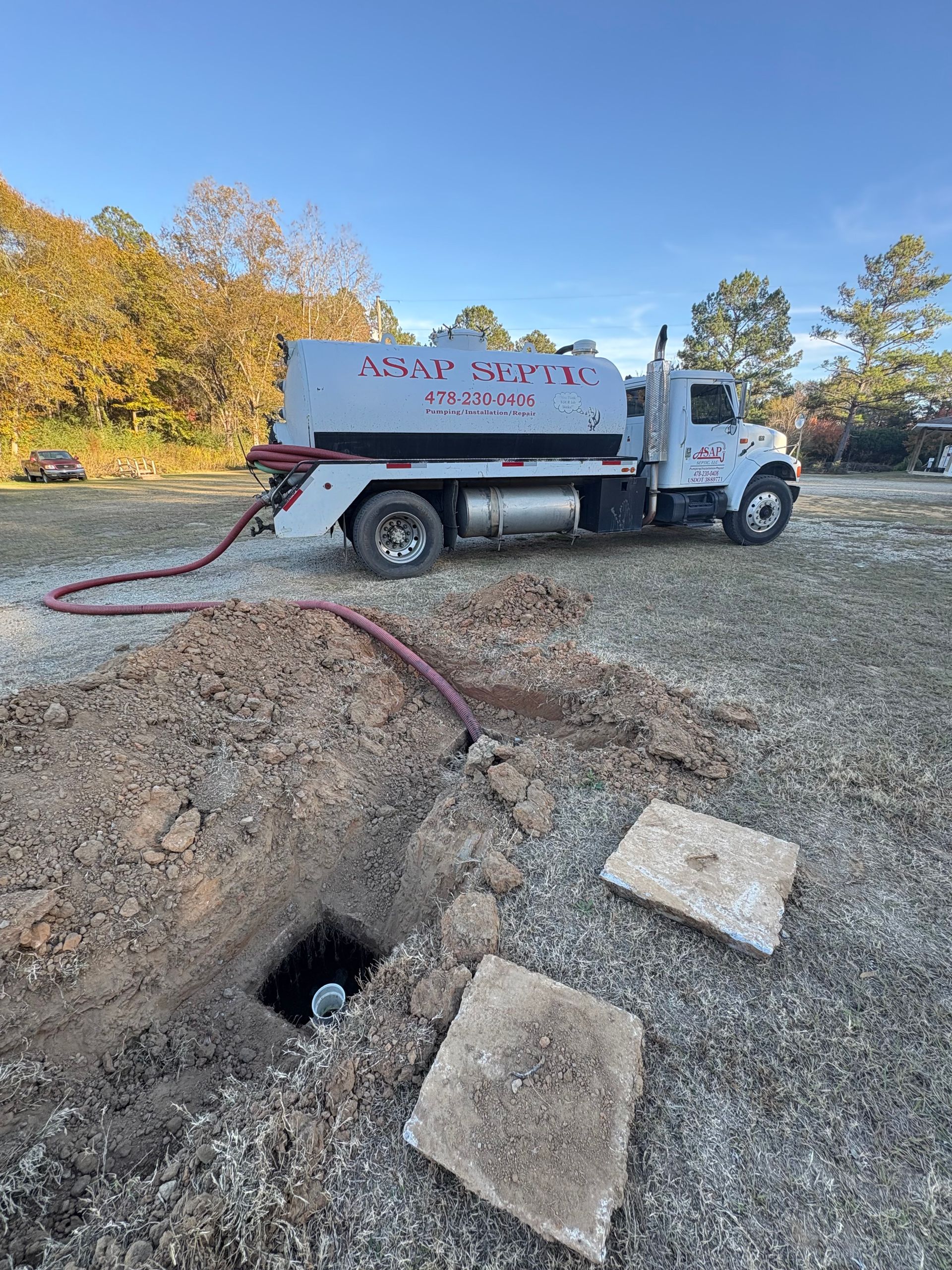 Construction site with a building and backhoe. Rows of gray drainage pipes lie on the ground, surrounded by mulch and trees under a blue sky.